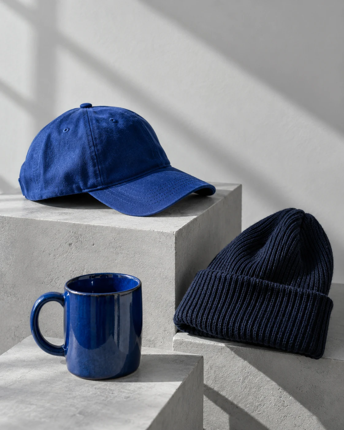 A blue baseball cap, navy knit beanie, and blue ceramic mug arranged on pale gray plinths
